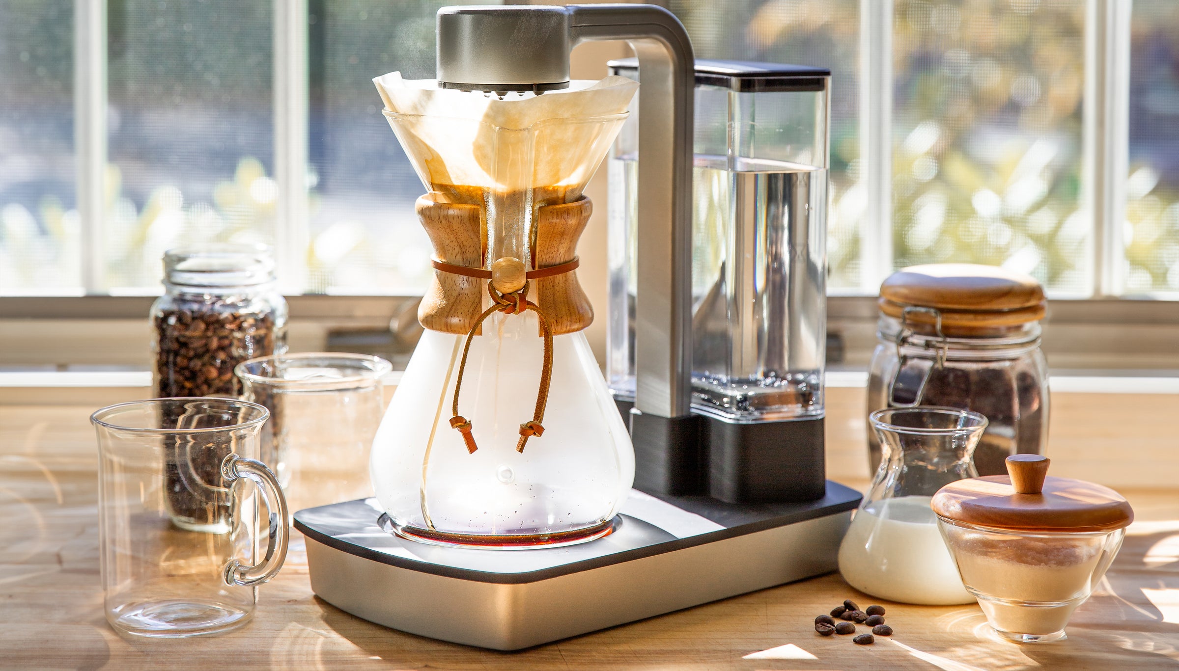 ottomatic brewing coffee on a kitchen counter in the morning sunshine. also shown 2 murray mugs and cream and sugar set and containers of coffee beans