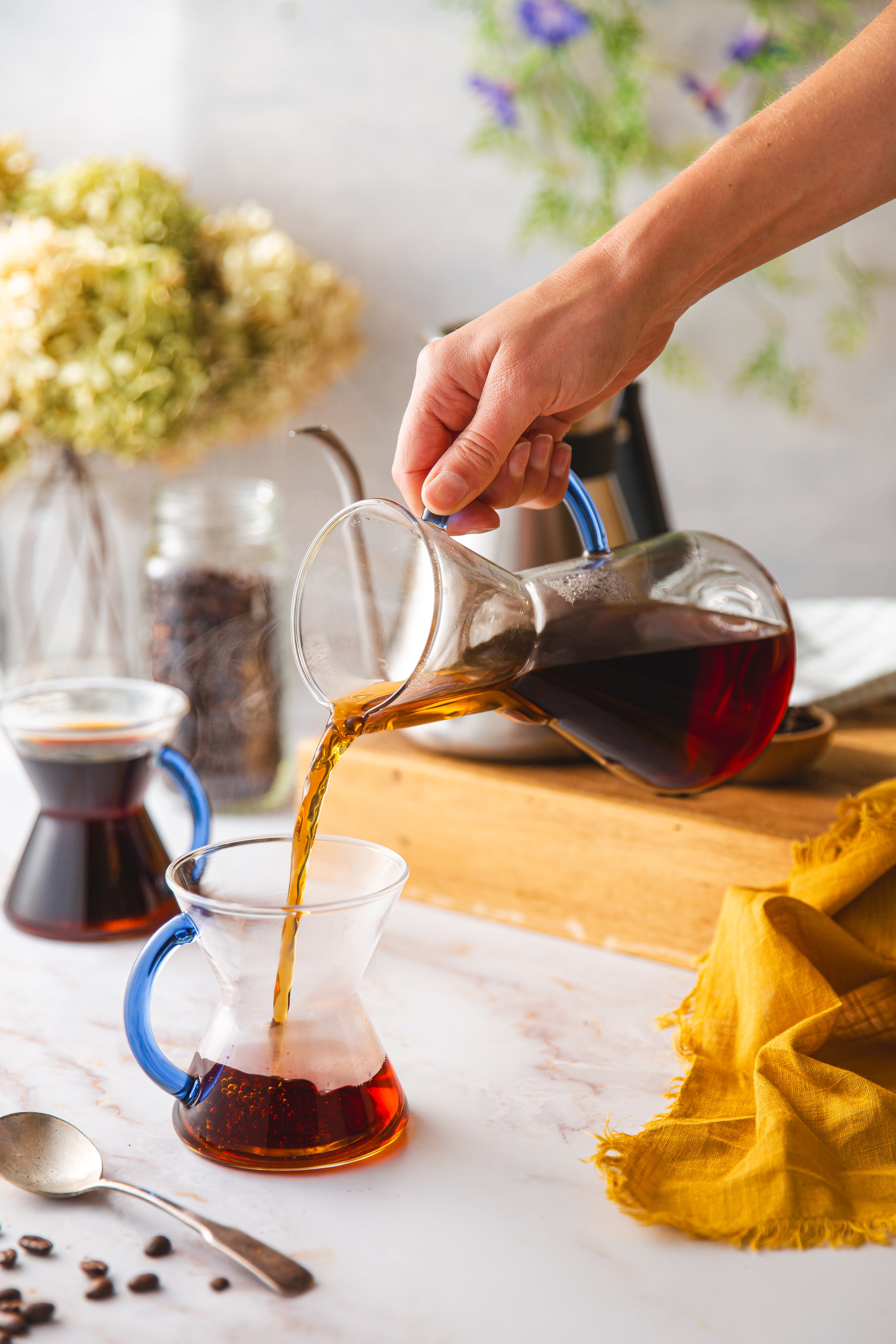 3-cup sapphire glass handle chemex pouring coffee into 1 of 2 sapphire glass handle mugs. sitting on a marble counter top with flowers, coffee beans and the chettle.