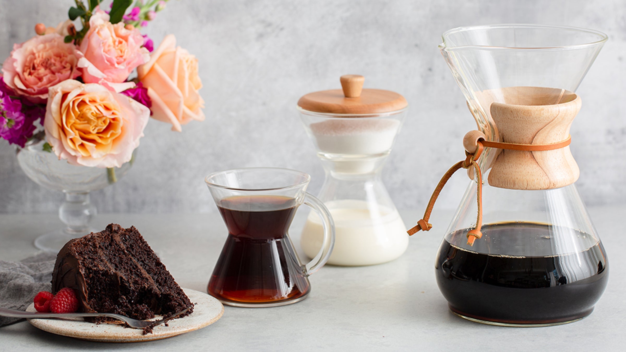 Coffeemaker, cream and sugar set and glass handle mug with a piece of chocolate cake and flowers 