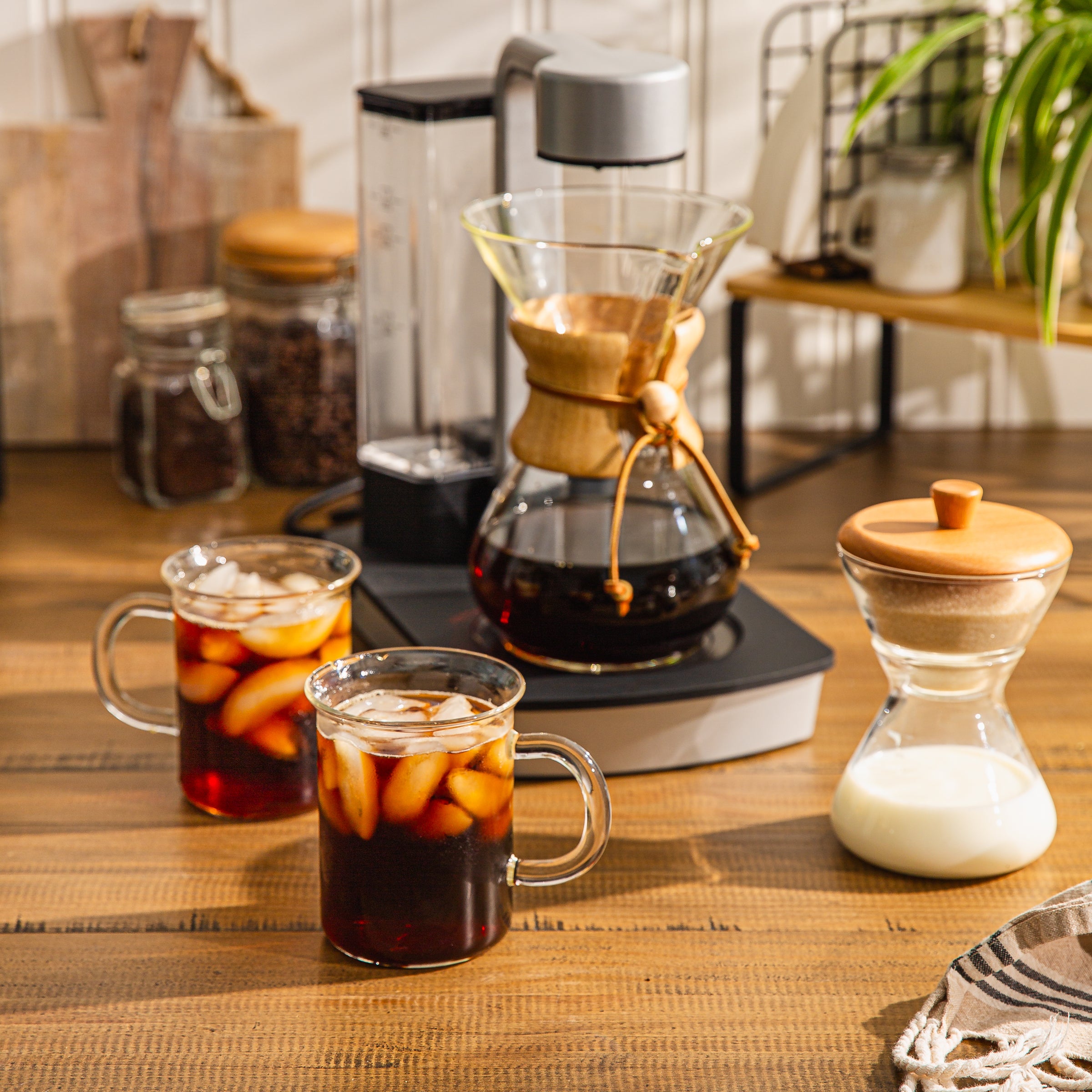 The Ottomatic Coffeemaker brewing iced coffee. Two Murray mugs filled with iced coffee, also on the wooden table is the Cream and Sugar set.