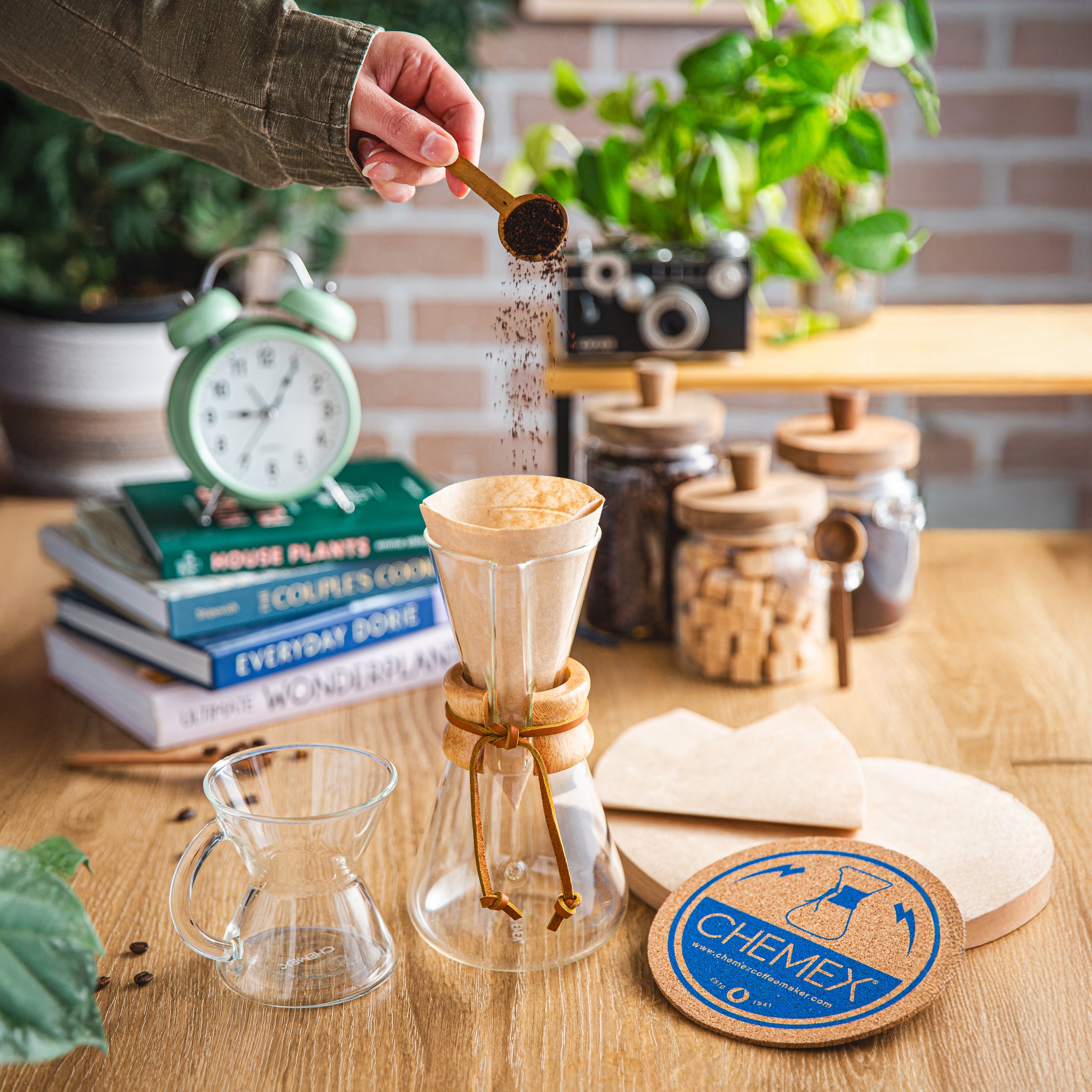 Person preparing coffee using a Chemex 3-cup Classic on a wooden table with books and plants in the background.