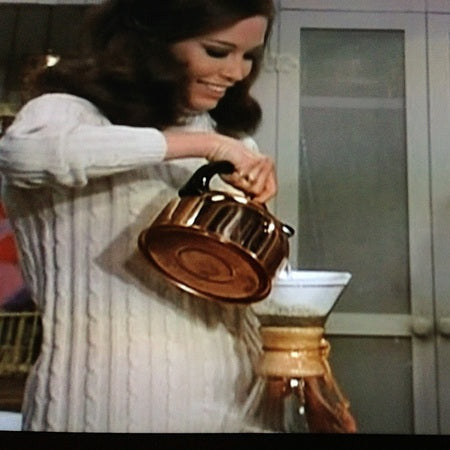 This image shows a scene from The Mary Tyler Moore Show featuring a person pouring water from a copper kettle into a distinctive Chemex glass coffee maker.
