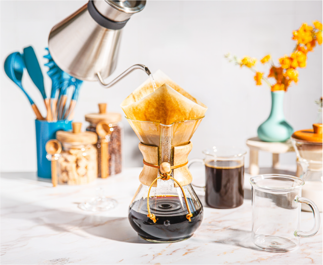 CHEMEX Classic Series coffeemaker mid-pour on a marble countertop — gooseneck kettle, brewed coffee, utensils and flowers, 2026