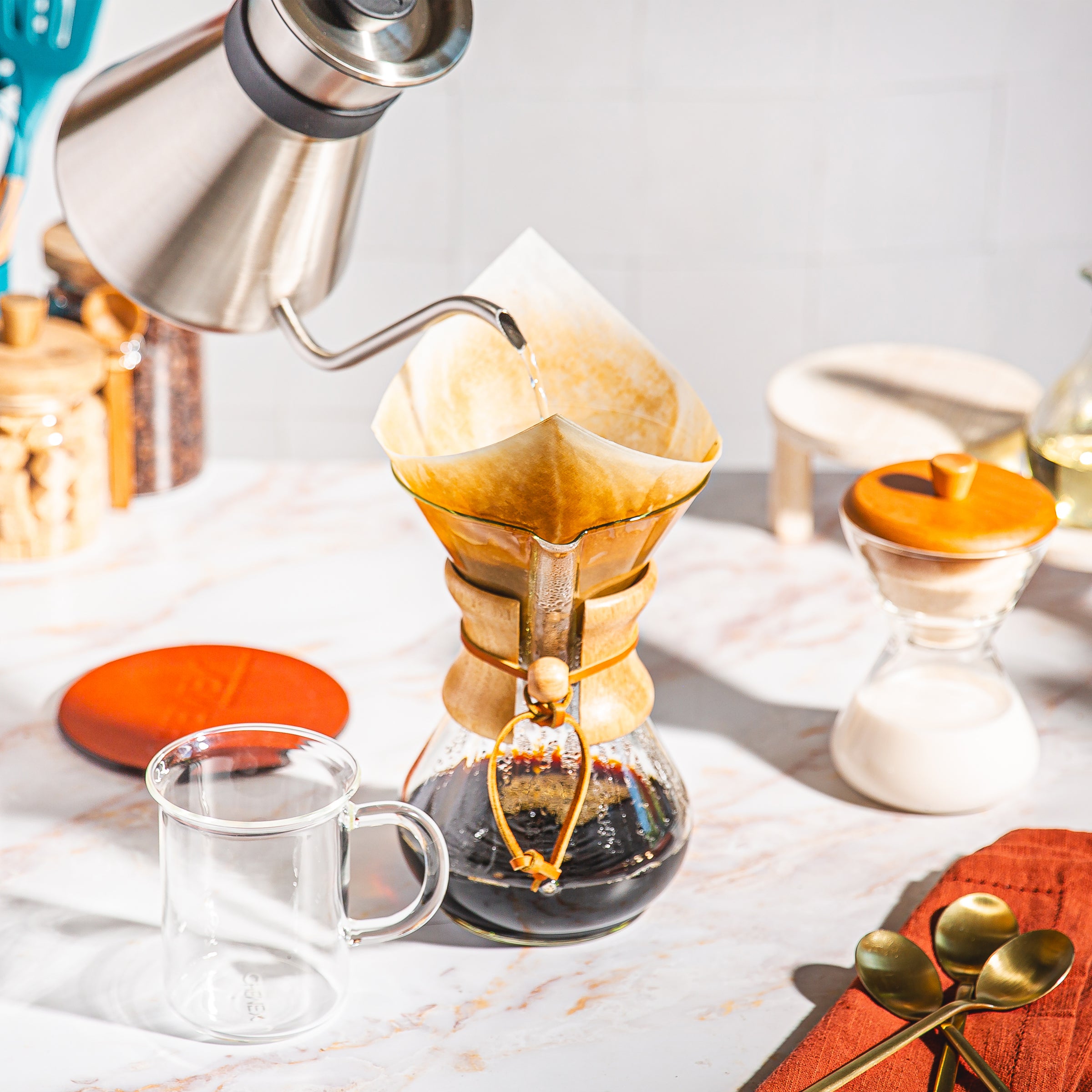 Hot water being poured from the chemex chettle into the six cup chemex pour-over, brewing delicious coffee. Murray Mug  sits empty on a marble surface in front of the coffeemaker. the cream and sugar set is also on the counter off to the right.